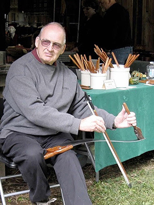 Stewart Walton sits on a folding chair beside a table of their wares at a craft fair.  He is holding a saw and drawing a fiddle bow across it with his right hand. In his left hand he is holding a handle attached to the far end of the saw blade.  He can make different notes by changing the position of that handle.