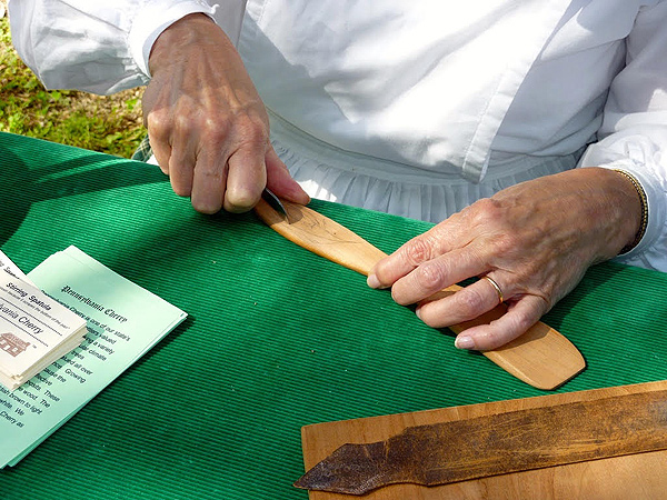 Closeup of Judy’s hands as she starts to carve a design in a spatula handle.  Her knife follows a pencil line draft on the wood. She is wearing a white long-sleeved dress in the Mennonite style.