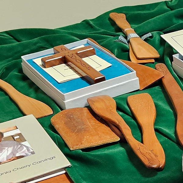 A display of items that the Waltons have made out of cherry wood,  decorative folk-art carvings. Laid out on a green velvet tablecloth are several stirring spatulas and a cross in a small box.