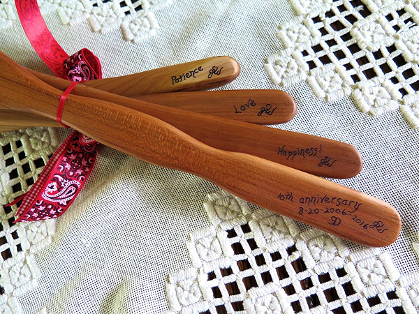 The same four spatulas as above, but flipped over to show the other side of the handle.  Each one has an inscription: Patience, Love, Happiness, and 10th Anniversary. They are laid on a handmade lace table runner.