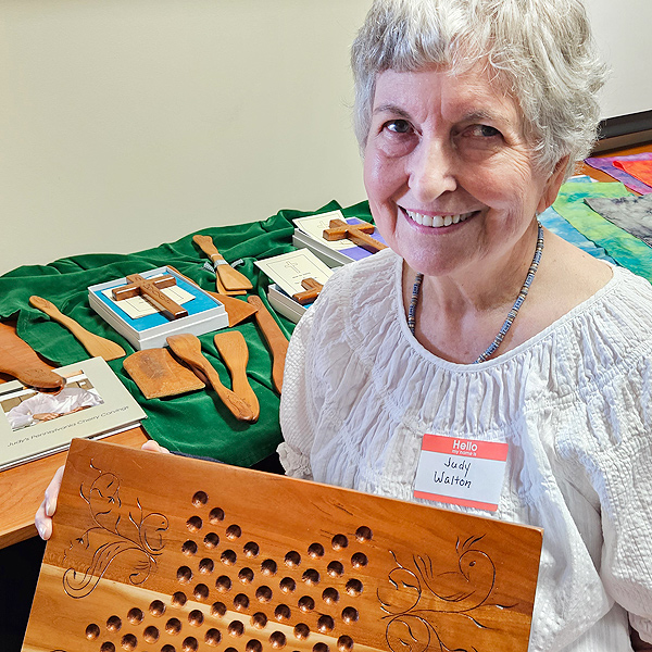 Judy Walton sits in front of a table holding her woodcarvings, holding up a Chinese Checkers board that she made, with Pennsylvania Dutch-style carvings in the corners. Judy appears to be in her 70s, with short gray hair and a warm smile.