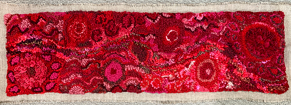 This hooked piece appears to be about one foot high and four feet wide. It’s an abstract design in intense reds, magentas and maroons. Evenly spaced on the piece are six round spots, concentric circles of red shades, each a few inches across. These are surrounded by abstract squiggles, curves and curlicues in various red shades.