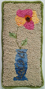 This wall hanging appears to be about 9 inches wide and 18 inches tall, and features a tall blue vase with a single large, colorful flower on a long stem. The design is more primitive than realistic. The flower has five large petals; three are done in shades of magenta, while two others (left and right) are done in shades of gold and peach.  The flower center is red, with dark stamens in the very center that are taller than the rest of the loops. The vase is also shaded, dark blue at bottom and fading as it goes up, with an abstract design of darker curlicues decorating it.  The background is solid tan. Overhand stitches of dark green yarn bind the edge.