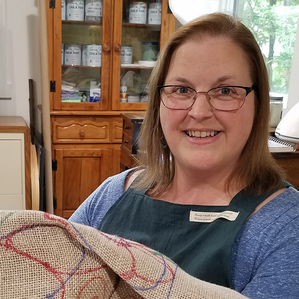 Julie Smith smiles for the camera, holding up a piece of linen rug backing with a design drawn on it.  She has an oval face, shoulder-length straight sandy hair, glasses, and a warm smile. She appears to be age 30-40. She’s wearing a dark blue apron over a heather-blue long-sleeved tee shirt. The design outlines on the backing are drawn in colored markers, and show large flower petals, flower stamens, and spirals that will be the background color.  Behind her are various cabinets holding supplies, and large windows looking out on her wooded yard.