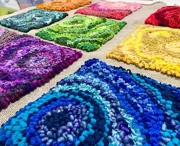 This work is made up of nine squares, each about eight inches across, spaced about an inch apart on the backing material. Each block is a different basic color (vibrant blue, violet, gold, green, red, etc) but is made up of many different shades of that color, arranged in concentric circles, curves, squiggles, etc. The view is angled, with the camera held close to the blue square at the bottom right, looking diagonally across the work to an orange square at the upper left.