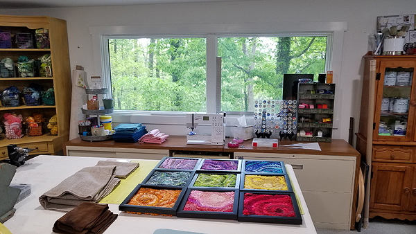 In the foreground is a worktable holding some folded pieces of wool fabric in earth tones, and a hooked piece that is nine squares within black wooden frames. Each of those squares is a different bright color, with lighter and darker tones in abstract patterns.  Behind the work table are various cabinets: tall hutches on either side of large windows and wood-topped wide filing cabinets under the windows. The windows look out on Julie’s wooded yard.  Colorful rug-making supplies are neatly displayed everywhere.