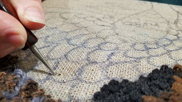 Closeup of Julie making a hooked rug.  Most of the frame shows the coarse linen backing material, beige with a pattern of overlapping ovals (short feathers) drawn in marker.  The bottom left and right corners are completed areas, with short loops of wool in shades of brown, blue and black. Julie’s right-hand finger and thumb are in the upper right corner, holding the hook down against the fabric.  A dark spot just beside the hook is really a widened gap between the threads of the weave; she has just demonstrated how to slip the hook through the fabric.