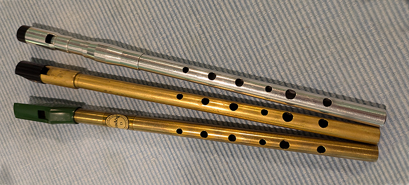 Three tin whistles are arranged in a fan, with their bottom ends touching.  These are metal tubes about a foot long and about 1cm in diameter.  Each has six finger-holes, some holes larger than others.  The bottom is brass with a green plastic mouthpiece. The middle is brass with a larger-diameter brass-and-Delrin mouthpiece. The top is shiny aluminum with a black Delrin tip on the mouthpiece.  The player blows into the mouthpiece, which directs the air across a square window an inch below the top of the mouthpiece.  The bottom edge of the window is filed to a sharp angle.