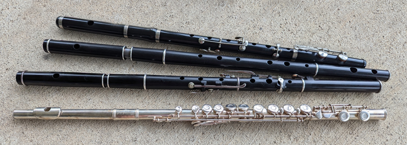 Four flutes are arranged in a fan, foot-joints close together and head-joints further apart. The bottom flute is shiny silver with more than a dozen keys and rods.  The others are black wood with silver rings at the joints and ends, and they all have six finger-holes, plus an embouchure hole in the head-joint which the player blows air across.  Two of the wooden flutes have a few silver keys, simple levers with a pad at one end to cover a hole; most keys are parallel to the flute body but a few go across it.