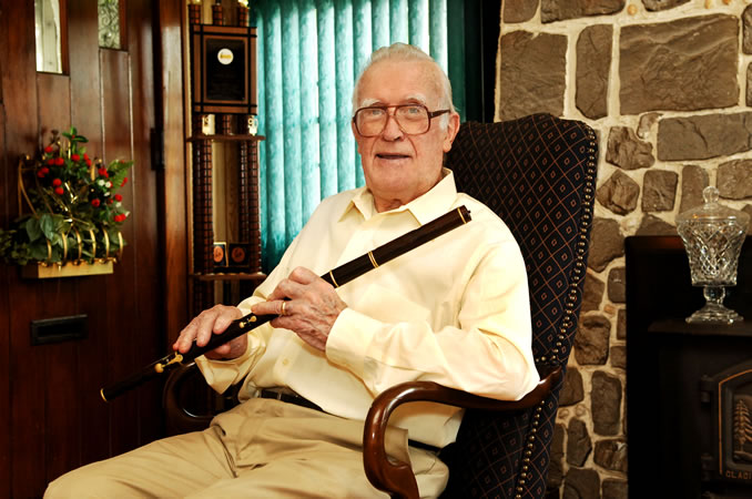 Mike Rafferty sits in a chair in his home, with a stone fireplace to his left and a wood-paneled wall to his right. He is an elderly man (age 83 in this photo) with white hair, bushy white eyebrows, and large squareish glasses with brown frames.  He is smiling slightly, as if about to speak. He wears a pale yellow dress shirt and khaki slacks.  Across his body he holds a wooden flute at an angle; it is black wood with gold rings and keys.