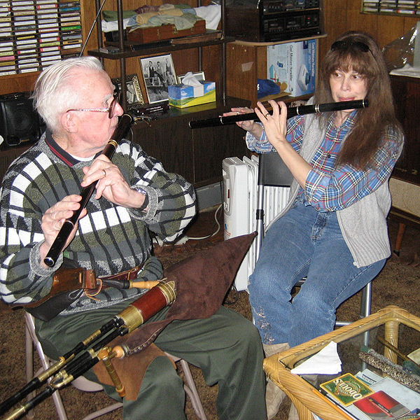 Mike Rafferty (at left) and Lesl Harker (at right) play flutes together in his basement.  They sit on folding chairs, and there are cassette tapes and photos and instrument cases on shelves behind them.  Their flutes are black wood with silver or brass rings, and have no keys. Mike is an elderly man who appears to be in his 80s, with white hair and glasses.  Lesl appears to be around age 40, casually dressed in flannel shirt and gray sweater vest.