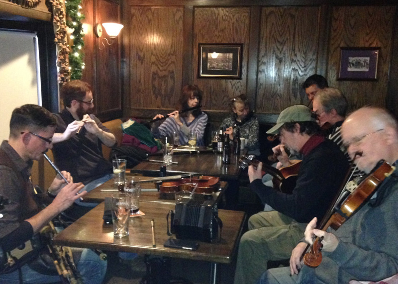 A group of eight musicians (two women, six men) play Irish music while sitting around a table in a pub.  There are flutes, tin whistles, fiddles and guitar being played, plus uilleann pipes and concertina not being played. All the players appear to be between ages 40 and 60. The walls are paneled in dark wood, and a garland of pine with tiny lights suggests the Christmas season.