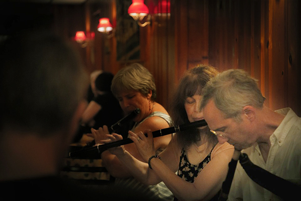 A dramatically-lit photo of people playing Irish music in a wood-paneled pub.  At the right edge of the photo is a man playing uilleann pipes; he is holding the chanter vertically and a bit of the bag is visible.  To his right, Lesl is playing her black wooden flute, wearing a summery sleeveless dress.  To her right, and almost in the center of the photo, is another woman playing flute, also in a sleeveless top, but she is almost all in shadow. At the left edge of the photo, but darkly shadowed, is the back of another musician’s head.