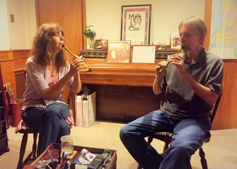 Lesl (at left) and her apprentice (at right) play whistles together.  They are seated in wooden chairs in her former home in New Jersey; she has her legs crossed.  He appears to be age 50 or so, with graying hair and a short beard. In the background is a piano with pictures and a plant on it, and an open door with a large map of Ireland pinned to it.