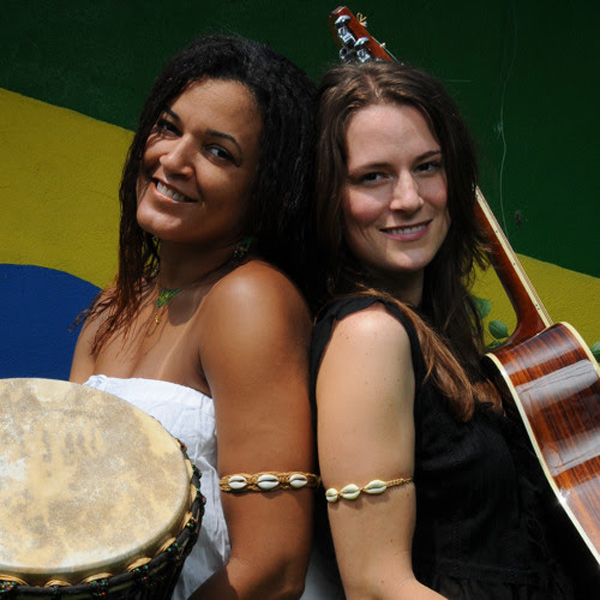 Monica Teles with her musical friend Chelsea Caroline.  In this promo shot for their duo Batida, they are back-to-back with their heads turned toward the camera.  Monica is holding a large Brazilian drum with a mottled leather drum-head; Chelsea is holding a guitar so that only its side and neck are visible.  Both wear an upper-arm band featuring three cowrie shells.  Monica has a lovely smile with a little mischief in it; she has shoulder-length black hair with a little crimp texture, and she’s wearing a white dress that shows off her bare shoulders and tawny brown skin. Chelsea has straight brown hair, a direct gaze, and fair skin.
