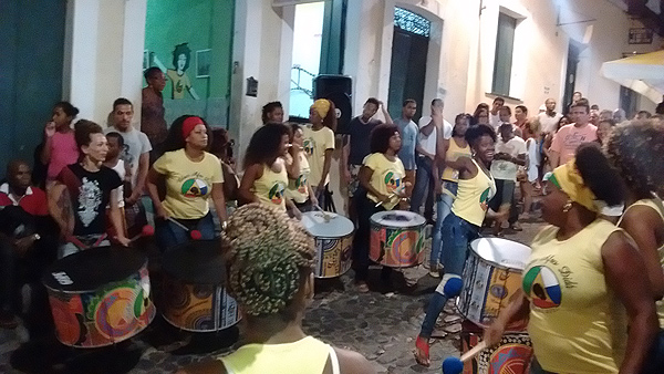 An alleyway in Salvador, Brazil, crowded with onlookers and several female percussionists with large colorful cylindrical drums.  The drummers are in a loose circle and a tall young woman is grinning and dancing in the open middle. The women are a range of ages and sizes; all are wearing yellow T-shirts with the school logo.  Nearly all of the onlookers and musicians are people of color. It appears to be night-time but the area is lit from above.