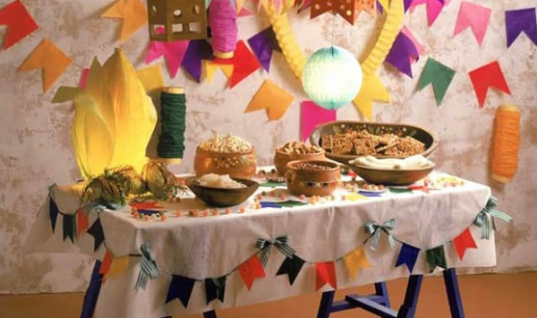 Here’s a table decorated for the Festival of St John celebration. The table is about 4 feet wide and 2 feet deep, with a simple white tablecloth, holding several large wooden and pottery bowls heaped with traditional foods. Pinned to the drape of the cloth is a garland string of brightly-colored little flags. On the wall behind the table are more garlands of larger flags, and other colorful decorations. At the back corner of the table is a glowing decoration made of paper, that looks a little like an ear of corn with the husks pulled back at the top.