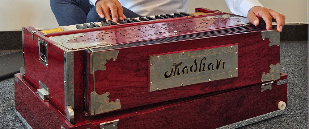 A close-in shot of the back of a harmonium, with reddish mahogany wood, engraved silver plates protecting the corners and edges, and a keyboard on the far side. A man’s hands are visible, one on the keyboard and one on the back panel.