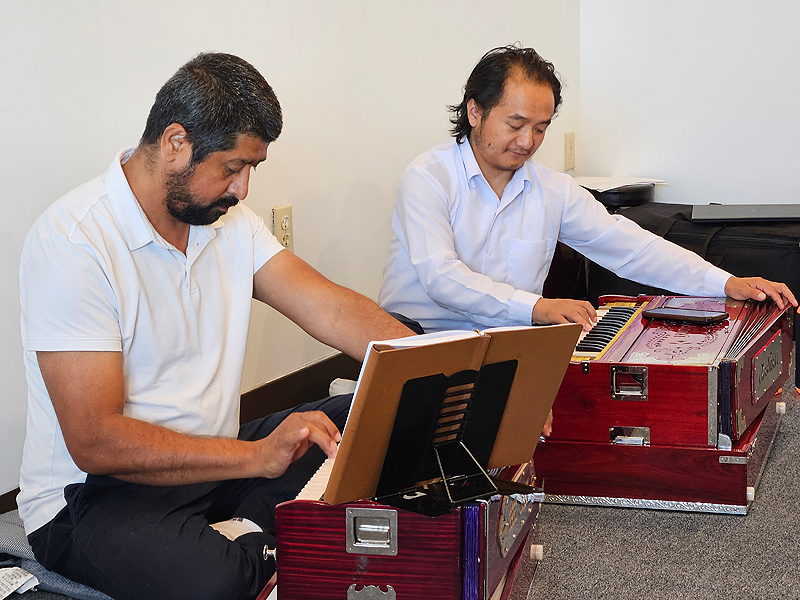 Two men sit on the floor, each of them concentrating on playing the harmonium in front of him.
