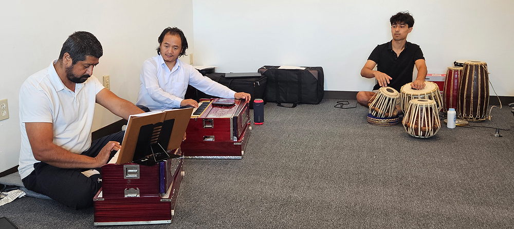 Two men sit on the floor, each with a harmonium. The student concentrates on his instrument; the teacher is explaining something.  Off to one side is a teenage boy with curly black hair, playing small hand drums that sit on the floor.