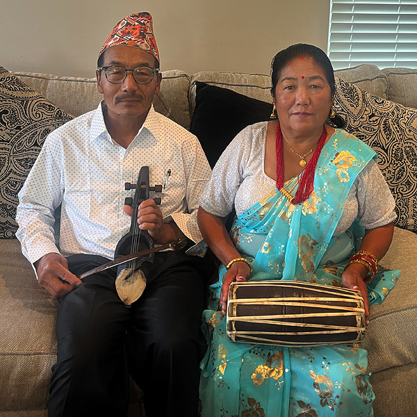 Two Nepali people sitting on a couch, holding instruments. They appear to be in their 60s. The man is holding a small stringed instrument with a bow. He is wearing a white dress shirt and a colorful almost-conical hat. The woman is holding a long skinny double-headed drum on her lap, with one hand on each drum head.  She is wearing a green silk sari and a tee shirt, with a multi-strand necklace of red beads.  She has dark hair, pulled back, and a small red bindi between her eyebrows.