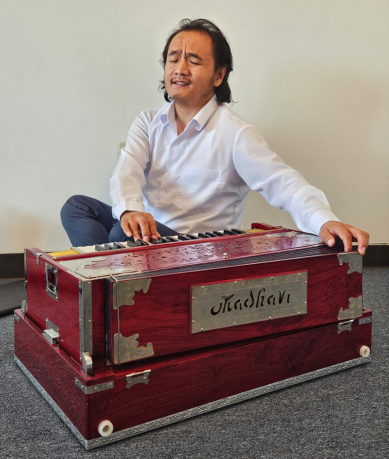 Muskan sits on the floor playing the harmonium, facing off to the side rather than facing the keyboard.  His left hand reaches across the instrument to work the bellows; his right is on the keyboard.  He is singing, with his eyes closed and a soulful expression.  He is a small, stocky man with shoulder-length black hair.