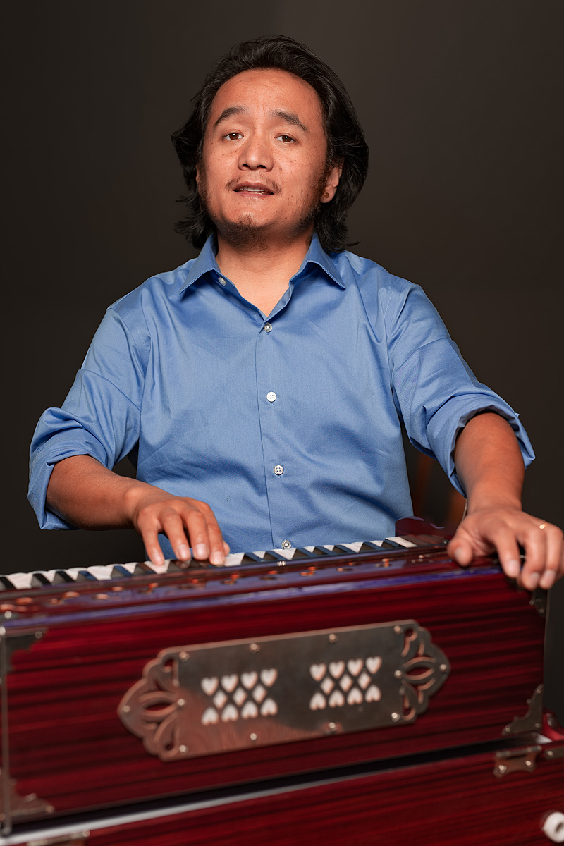 A studio photo of Muskan Balampaki playing the harmonium and singing.  He has his right hand on the keyboard, and his left extended over the instrument to work the bellows at the back. He is a small, stocky Nepali man with an oval face, shoulder-length black hair, scanty moustache and goatee.