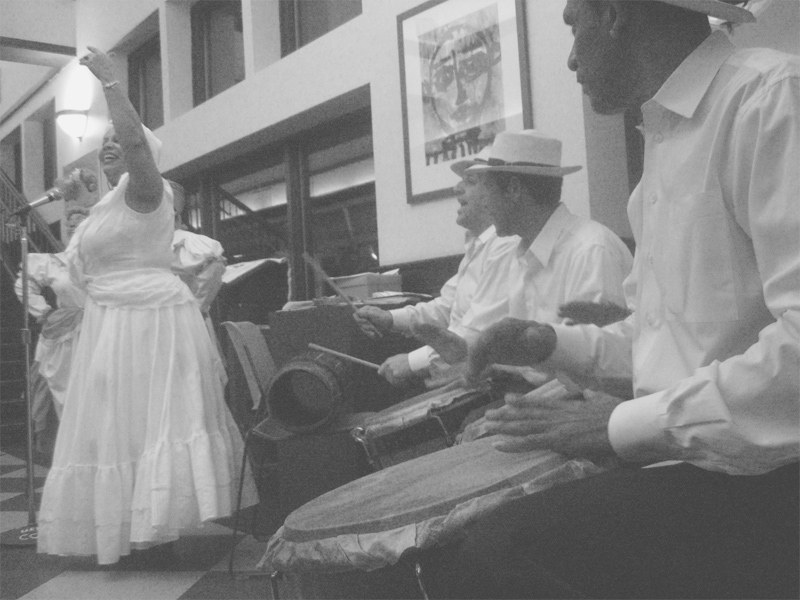 This image from Wikimedia shows a performance of a bomba band in what appears to be a building lobby.  There are three drummers to the right, all wearing white dress shirts and white fedora hats. At the left side of the image is a large woman in a long, frilly white dress, gesturing upward with her left arm raised, and grinning.