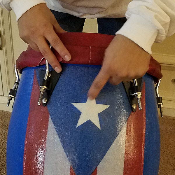 Pedro shows a drum that he made.  The body is painted with a Puerto Rican flag; he is pointing to a white star within a blue triangle. Below the triangle are alternating red and white vertical stripes.  The drum is about 16 inches in diameter and 3 feet tall, and has a red leather drum-head attached with metal brackets.