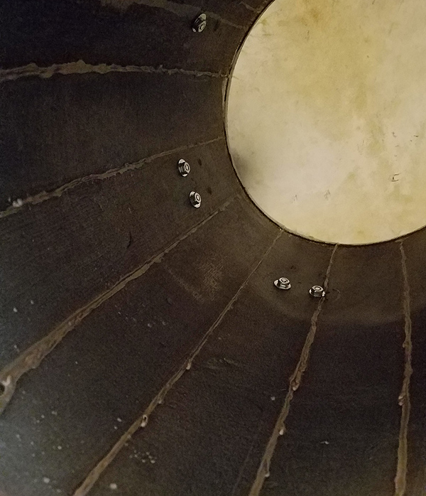 Looking up into a bomba drum from below.  The head of the drum appears as a round white circle at the top right of the image, and the barrel-like staves of the drum body are dark wood with lighter-colored sealant, which makes them look like sun rays. A few inches down from the head are bolts which secure the drum-head tuners to the outside.