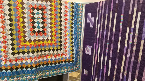 Two quilts side by side.  The one on the left has concentric rectangles of colored blocks, rotated to the diagonal so blocks of the same color meet only at their points.  A column of four blue calico blocks in the center, surrounded by ten light-colored blocks, surrounded by black, then red/gold print, then blue, then white/pastels, then brown, and so on out to the border.  The quilt on the right has thin vertical strips of different prints in shades of lavender. Its background is purple, and though the picture doesn’t show it, the purple is glittery.  The border, visible at the center of the photo, is mostly purple with lavender ’sampler blocks’ showing different traditional quilt patterns: drunkard’s path, grandmother’s fan, shadow box.