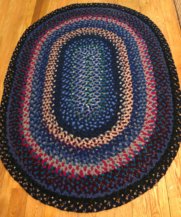 A braided wool rug on a hardwood floor; the rug appears to be about 5ft x 7ft. The concentric ovals are each about three braids wide and feature several shades of blue accented with red, maroon, gray, black, a little green, and various plaids.