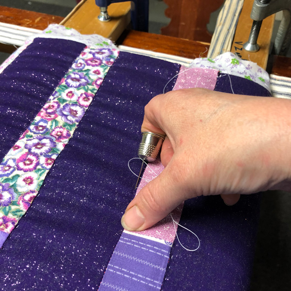 Close-up of a woman’s right hand putting a quilting stitch into the purple quilt. At the top of the shot are the clamps holding two long pieces of wood around which the quilt top is wrapped.  A thimble is on the quilter’s index finger, and she’s using it to push the needle down into the fabric layers at an angle. Her thumb is resting on the fabric.