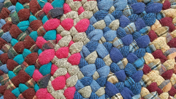 A closeup of the ‘Rhapsody in Blue’ braided rug, showing about twelve rows of braids, illustrating the range of colors that went into it.  From left: maroon, medium blue, teal, and a plaid; then magenta and gray tweed; then light & medium blue with solid gray; then dark & medium blue with blue plaid; then maroon & two tan plaids.
