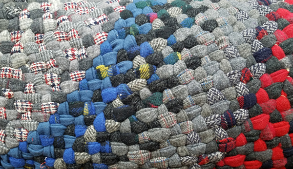 Another closeup of a braided rug, this one showing more of the rug, which looks mostly gray (lots of plaids in the braids) with a band of blue and a band of red.