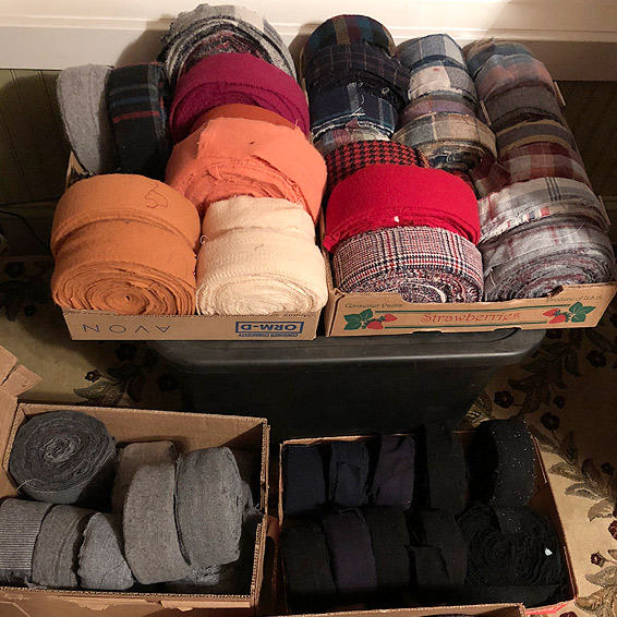 Four shallow cardboard boxes hold rolls of wool strips set up on edge, sorted by color.  At top, rolls of peach, tan, cream, red, and various plaids; at bottom, a box of gray and a box of black.