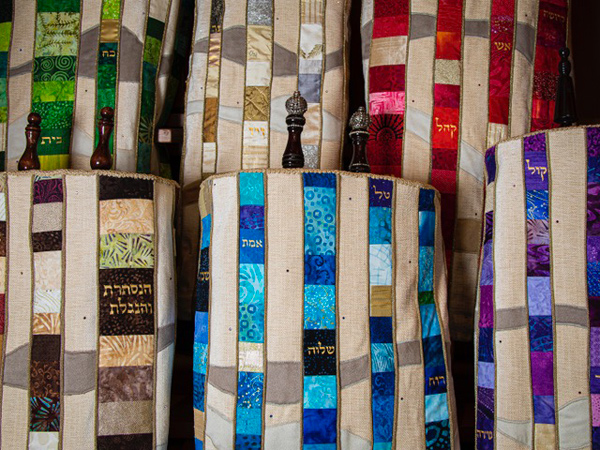 The Torah scrolls themselves appear to be about 30 inches tall, and are stored vertically.  The mantles are quilted in stripes: alternating colored strips (some with Hebrew writing) and lighter beige strips.  The design echoes the sculptural cabinet that houses the Ark.