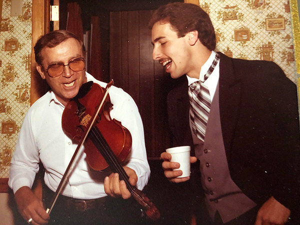 George Caba is a middle-aged man, somewhat stout, wearing a white buttoned shirt and large aviator-style glasses. He is playing fiddle and singing.  Lenny Tepsich is at right, singing with his eyes closed. He’s wearing a black tuxedo jacket, white wing-collar shirt, a brown vest, and a brown-and-white striped necktie that’s much wider than a standard tie. The feel of the photo suggests the 1970s.