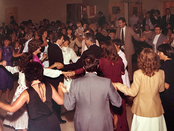 Kolo dancers pack a large room at a church, smiling and looking like they’re having fun.  They are all wearing dressy clothes (they are at a wedding reception) and holding hands in lines or circles.  Clothes and hairstyles suggest the 1970s.