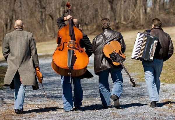 The four members of Sviraj walking away from the camera, on a flat gravel road, with their instruments on their backs.  They are wearing winter coats, and the vegatation in the background is leafless.