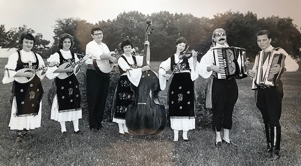 Black-and-white photo of a tambura band, all wearing traditional Serbo-Croatian clothing.  The four women wear dark embroidered pinafores over white dresses; the three men wear white shirts and black pants. From left to right, there are three players of stringed instruments; a woman with upright bass in the middle; a fiddler; a man with an accordion, wearing a head scarf, sash, and pants that show his ankles. On the far right is teenage Danilo with his accordion, pants tucked into shiny knee-length boots.