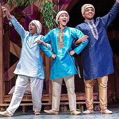 Three boys gesture dramatically, radiating excited happiness. They have their arms linked, and the boys on the outside are pointing upward with their free arms. They are wearing traditional garments of India: silk tunics and trousers, turbans, and silk slippers. The youngest boy is wearing pale blue, the eldest is wearing dark blue, and the middle boy is wearing bright medium blue. Their skin is light to dark brown.
