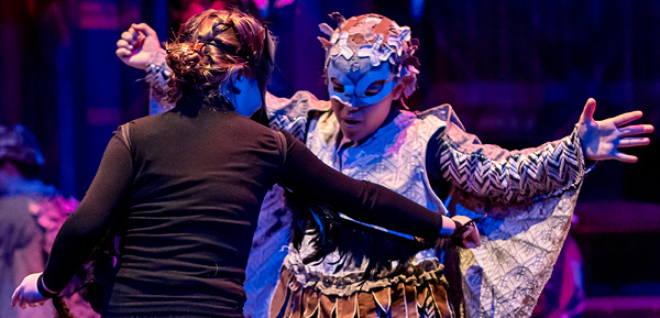 This still photo from the Panchatantra Tales production depicts the battle of the Crows and the Owls. Facing us is a dancer dressed as an owl, holding her arms out to show fabric wings hung from her arms. Facing her is a dancer wearing black, who seems to be attacking the owl with a sword. The owl dancer is looking down to where she's being struck in the belly.
