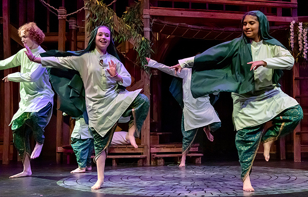 In this still photo from the Panchatantra Tales production, several barefoot young actors dance in a circle. They hold their left foot high, their right arm out to the left, and their left arm across their chest also pointing left. They wear traditional Indian tunics of pale green silk, with loose trousers and flowing head scarves of dark green.