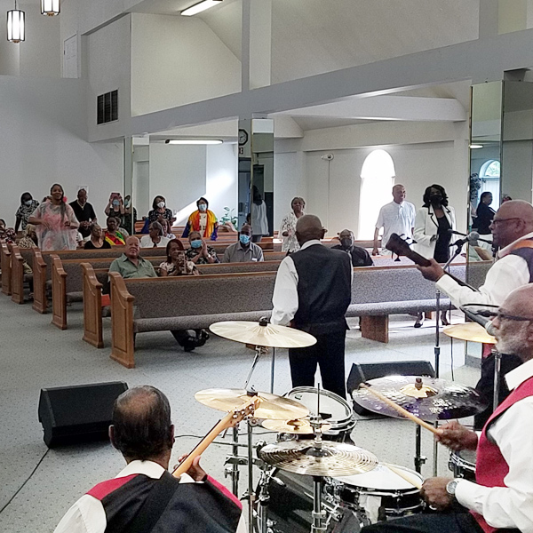 The Spiritual Messengers perform in a church. The photo is taken from behind the band and shows two vocalists out front, drummer and bass behind them, and audience sitting and standing in the pews.