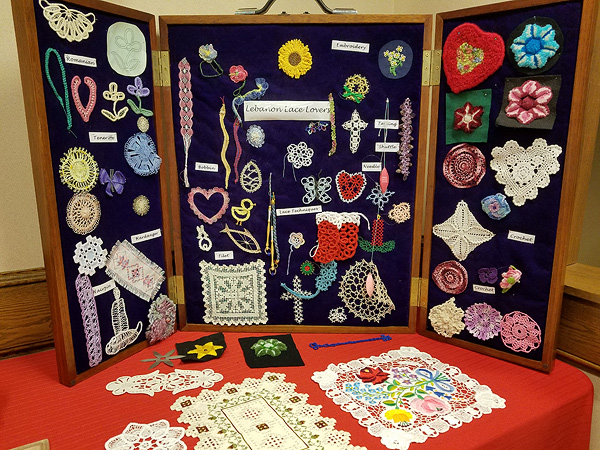 A wide variety of lace samples, pinned into a 3-panel display box that sits vertically on a table.  More lace samples sit in front of the box, laid on a red tablecloth. Several pieces are done in color, ranging from pastel doilies to vivid red Christmas stockings.  Several also include embroidery. Paper labels give the names of the various styles: Romanian, Teneriffe, Hardanger, Hairpin, Bobbin, Filet, Tatting (with a shuttle and needle) and Crochet.