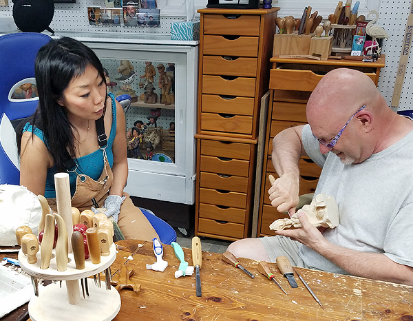 Aron Rook and Jim Hiser sit together behind a work-table. Aron is a young woman of Asian appearance, with straight black hair. Jim is a portly white man in his 60s, with a shaved head and a short white goatee. He is working on a wood-carving held in his lap.