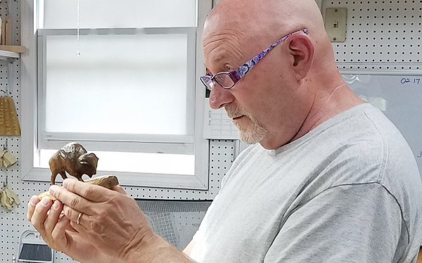 Jim Hiser, seen in profile, holds a small figurine of a buffalo in both hands, while looking over the tops of his purple-framed reading glasses at his apprentice who is not seen. 