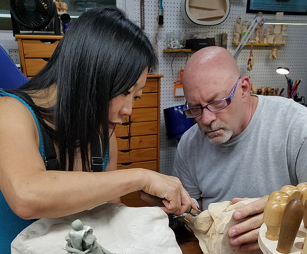 Aron Rook and Jim Hiser sit close together to work on a wood-carving. Aron is a young woman of Asian appearance, with straight black hair falling past her face.  Jim is a portly man of middle age with a shaved head and short white goatee.  He wears purple-framed reading glasses.