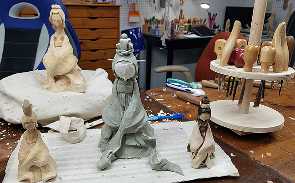 Three models of 'the young bride' character.  The largest and most elaborate, in the center, is made of gray clay, with flowing clothing and a complicated ornament on its head, but no facial features. At right is a smaller model in earth-tone colors, possibly a display piece in its own right. At left is a wood model, possibly a first draft. Behind those is Aron's current wood carving, nearly the same size as the largest model. 